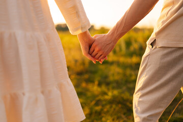 Wall Mural - close-up of unrecognizable young couple in love holding hands looking together to green field on sun