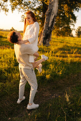 Wall Mural - Vertical shot of loving young man holding beautiful red-haired girlfriend in arms and looking into her eyes on green meadow in sunny summer evening, during golden sunset with soft sunlight.