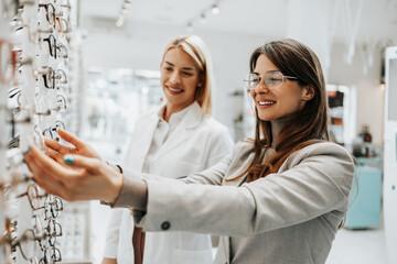 Sticker - Beautiful and fashionable woman choosing eyeglasses frame in modern optical store. Female seller specialist helps her to make right decision.