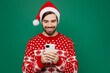© ViDi Studio - Merry cheerful happy fun young man 20s wears red warm knitted sweater Santa hat posing use mobile cell phone isolated on plain dark green background studio. New Year 2023 holiday celebration concept.