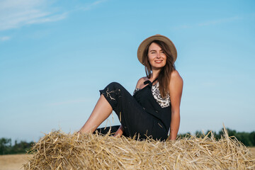 beautiful brunette woman in black overalls with a straw hat on a haystack in a field at sunset. High quality photo