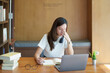 A portrait of a young Asian woman using a computer, wearing headphones and using a notebook to study online shows boredom and pain from video conferencing on a wooden desk in library