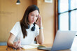 © Jirapong - A portrait of a young Asian woman using a computer, wearing headphones and using a notebook to study online shows boredom and pain from video conferencing on a wooden desk in library