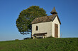 Eine kleine idyllische Kapelle auf einer grünen bergigen Wiese, mit einem herbstlichen Laubbaum und einer kleinen Bank, bei strahlend blauem Himmel.