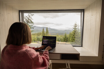 Woman works on laptop while sitting by the table in front of panoramic window with great view on mountains. Remote work and escaping to nature concept