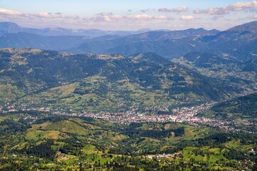 Poster - Aerial breathtaking view of small town in green valley surrounded by mountains under cloudy blue sky