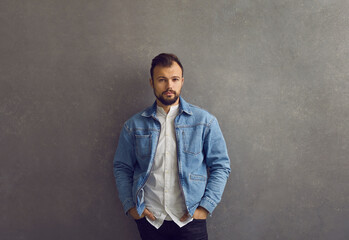 Handsome male fashion model posing against grey studio wall. Unsmiling young man in blue denim jacket and casual white shirt standing hands in pockets looking at camera with confident face expression