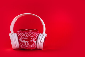 Christmas mood and vibe, warming traditional new year songs music listen concept. Red mug with christmas knitted sweater with big headphones on red background, minimal simple design copy space