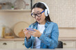 © Liubomir - Young beautiful Asian woman listening to music on the phone. Sitting at home in the kitchen wearing white headphones, holding a smartphone, scrolling through the playlist, choosing a song, smiling