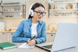 © Liubomir - Online training. Young beautiful Asian girl sitting at the table at home in white headphones and with a laptop. He studies remotely. Writes down a lecture, class in a notebook.