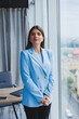 © DSMT - Portrait of a pretty brunette in a blue jacket in the office near the panoramic window with a view of the city. Happy business woman in the office