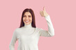 Beautiful positive Caucasian woman showing thumbs up and smiling broadly agreeing to promotional offer demonstrating approval dressed in white casual turtleneck stands on plain pink background