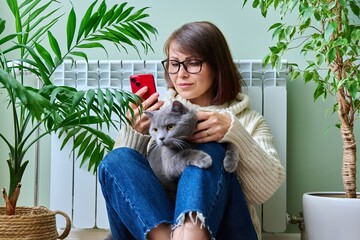 Wall Mural - Woman at home using smartphone, warming with cat near heating radiator