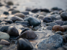 Pebbles On The Seashore Close-up. Rocky Beach. Stones Close-up With Bokeh. Gray Natural Background. The Concept Of Rest On The Seashore.  Autumn On The Seashore. Waves And Wet Pebbles