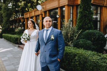 An adult stylish groom in a blue suit and a beautiful smiling bride in a blue dress are walking along the city street in the park, holding hands. Wedding photography of the newlyweds, portrait.