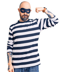 Poster - Young handsome man wearing burglar mask strong person showing arm muscle, confident and proud of power