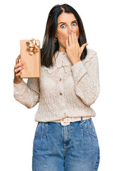 Canvas Print - Middle age brunette woman holding gift covering mouth with hand, shocked and afraid for mistake. surprised expression