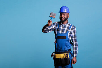 Professional house painter paints new house holds paint brush wears protective helmet and coveralls. Repair man happy doing interior renovation isolated on blue background.