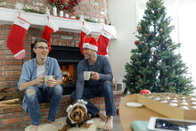 Smiling Father And Son Drinking Coffee With Dog Wearing Reindeer Antlers In Christmas Living Room