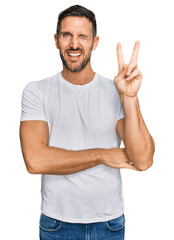 Canvas Print - Handsome man with beard wearing casual white t shirt smiling with happy face winking at the camera doing victory sign. number two.