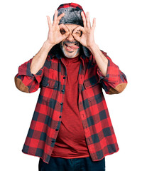 Poster - Middle age hispanic man with grey hair wearing fluffy earmuff hat doing ok gesture like binoculars sticking tongue out, eyes looking through fingers. crazy expression.