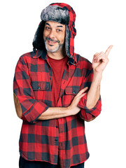 Poster - Middle age hispanic man with grey hair wearing fluffy earmuff hat with a big smile on face, pointing with hand and finger to the side looking at the camera.