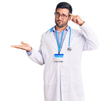 Young Hispanic Man Wearing Doctor Uniform And Stethoscope Confused And Annoyed With Open Palm Showing Copy Space And Pointing Finger To Forehead. Think About It.