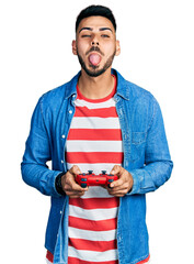 Wall Mural - Young hispanic man with beard playing video game holding controller sticking tongue out happy with funny expression.