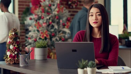 Poster - Portrait of asian employee working on laptop during christmas eve, using computer to create report. Sitting at desk filled with xmas decorations and festive ornaments, winter celebration.