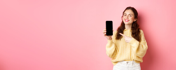 Wall Mural - Portrait of attractive young woman demonstrates promo on phone, pointing finger at smartphone and smiling, standing against pink background