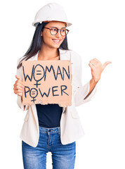 Poster - Young beautiful latin girl wearing architect hardhat holding woman power banner pointing thumb up to the side smiling happy with open mouth