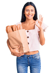 Wall Mural - Young beautiful latin girl holding paper bag with bread doing ok sign with fingers, smiling friendly gesturing excellent symbol