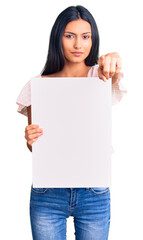 Canvas Print - Young beautiful latin girl holding blank empty banner pointing with finger to the camera and to you, confident gesture looking serious