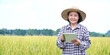 Portrait elderly asian woman wearing hat, holding mobile taplet and standing in rice paddy field, soft and selective focus, concept for smart farmer and happy senior woman in her own lifestlye