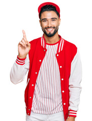 Canvas Print - Young man with beard wearing baseball uniform showing and pointing up with finger number one while smiling confident and happy.