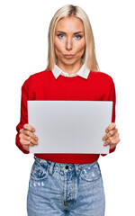 Wall Mural - Young blonde woman holding blank empty banner thinking attitude and sober expression looking self confident