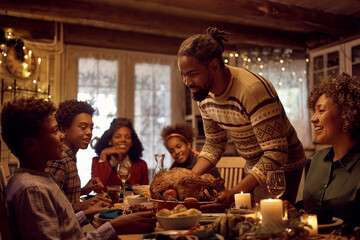 Happy black father serving Thanksgiving turkey to his family at dining table.