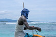 A Melanesian man sitting on the bow of a fishing boat wearing head wrap scarf with a large feather to protect from sun in Bougainville, Papua New Guinea