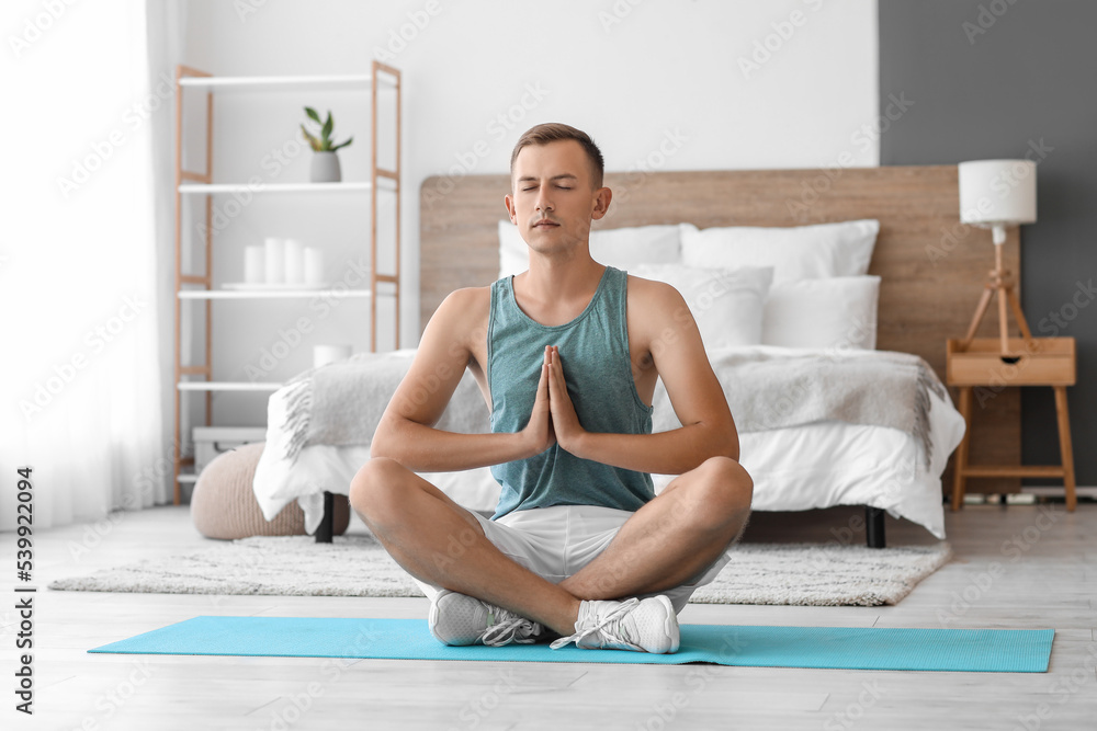 Young man meditating on mat in bedroom