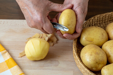 Wall Mural - Hands peel potato, peelings on wooden cutting board.