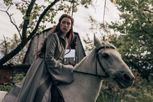 Young Woman In Image Of Ancient Rider Warrior Sits On White Horse In Cloak.