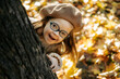 Cute little girl with a health disorder in a knitted sweater and hat and stylish glasses peeks out from behind a tree, plays hide and seek, has fun, enjoying the warm weather in a bright autumn park