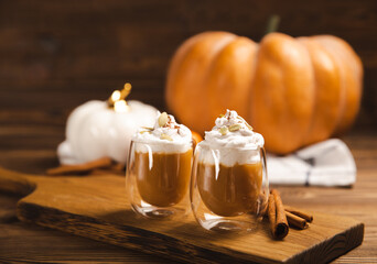 Pumpkin spice latte in glass mug with cinnamon, nutmeg, pumpkin seeds and whipped cream on brown texture wood. A glass of autumn coffee with pumpkin. A warming drink. Autumn concept.