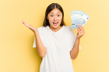 Wall Mural - Young asian woman holding a banknotes isolated on yellow background receiving a pleasant surprise, excited and raising hands.