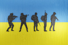 Paper Cutting Of Soldiers In Silhouette On Wooden Board Printed With The National Flag Of Ukraine. Illustration Of The Concept Of Ukrainian Armed Forces