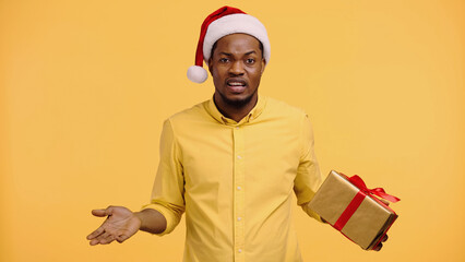 Canvas Print - confused african american man in santa hat holding present and showing shrug gesture isolated on yellow.