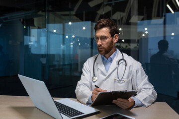 Mature doctor working on paper work inside a shabby office building, man in medical coat using laptop filling out medical documents concentrating and thinking