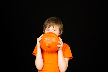 Wall Mural - a smiling boy in an orange t-shirt holds a pumpkin in his hands, a portrait of a boy on a black background