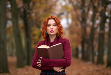 Wall Mural - Beautiful girl with red hair in a sweater reads a book in the autumn park, October