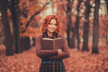 Wall Mural - Beautiful girl with red hair in a sweater reads a book in the autumn park, October